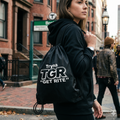 Person wearing a black hoodie with 'Tryna TGR Get Rite' text on a backpack, walking down a city street.
